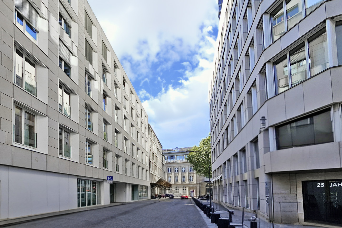 Straßenansicht zwischen zwei modernen Bürogebäuden mit hellen Fassaden und vielen Fenstern, im Hintergrund ein historisches Gebäude und blauer Himmel mit Wolken.