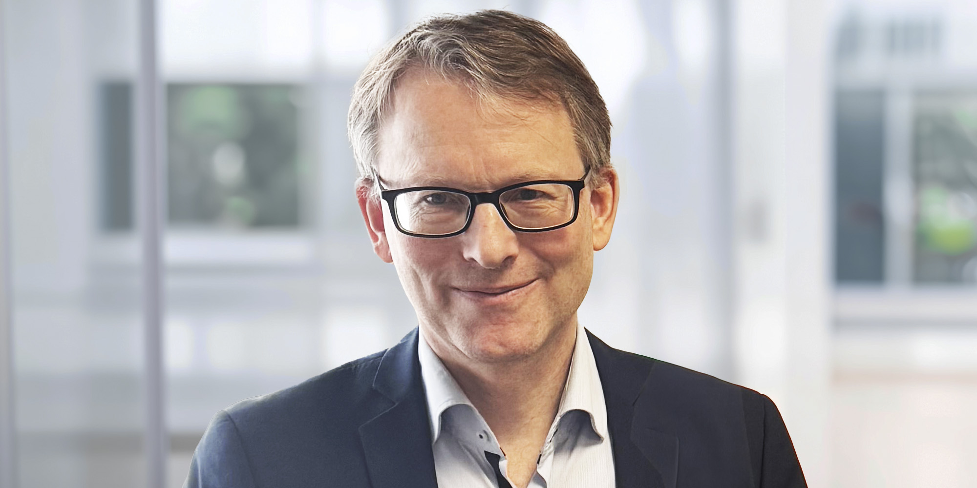 Michael Witt, lächelnd mit Brille und kurzem braunem Haar, trägt ein dunkles Sakko und hellblaues Hemd vor einem hellen, unscharfen Hintergrund.