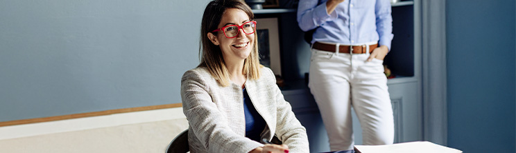 Lachende Frau mit roter Brille sitzt an einem Schreibtisch, im Hintergrund steht eine weitere Person – Symbol für moderne Führung, Kommunikation und Teamarbeit.