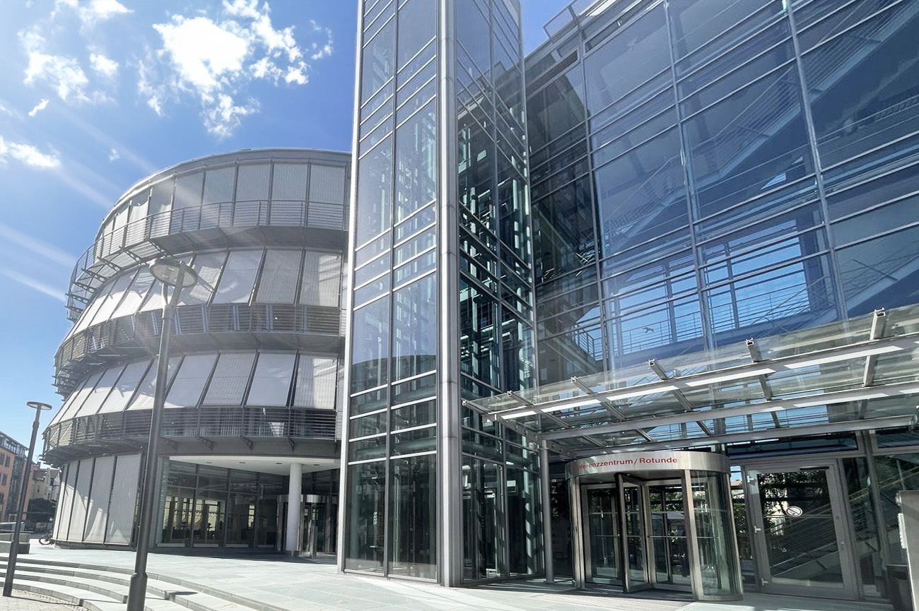 Moderne Glasfassade eines Bürogebäudes mit Eingangsbereich und rundem Anbau, bei sonnigem Wetter mit blauem Himmel.