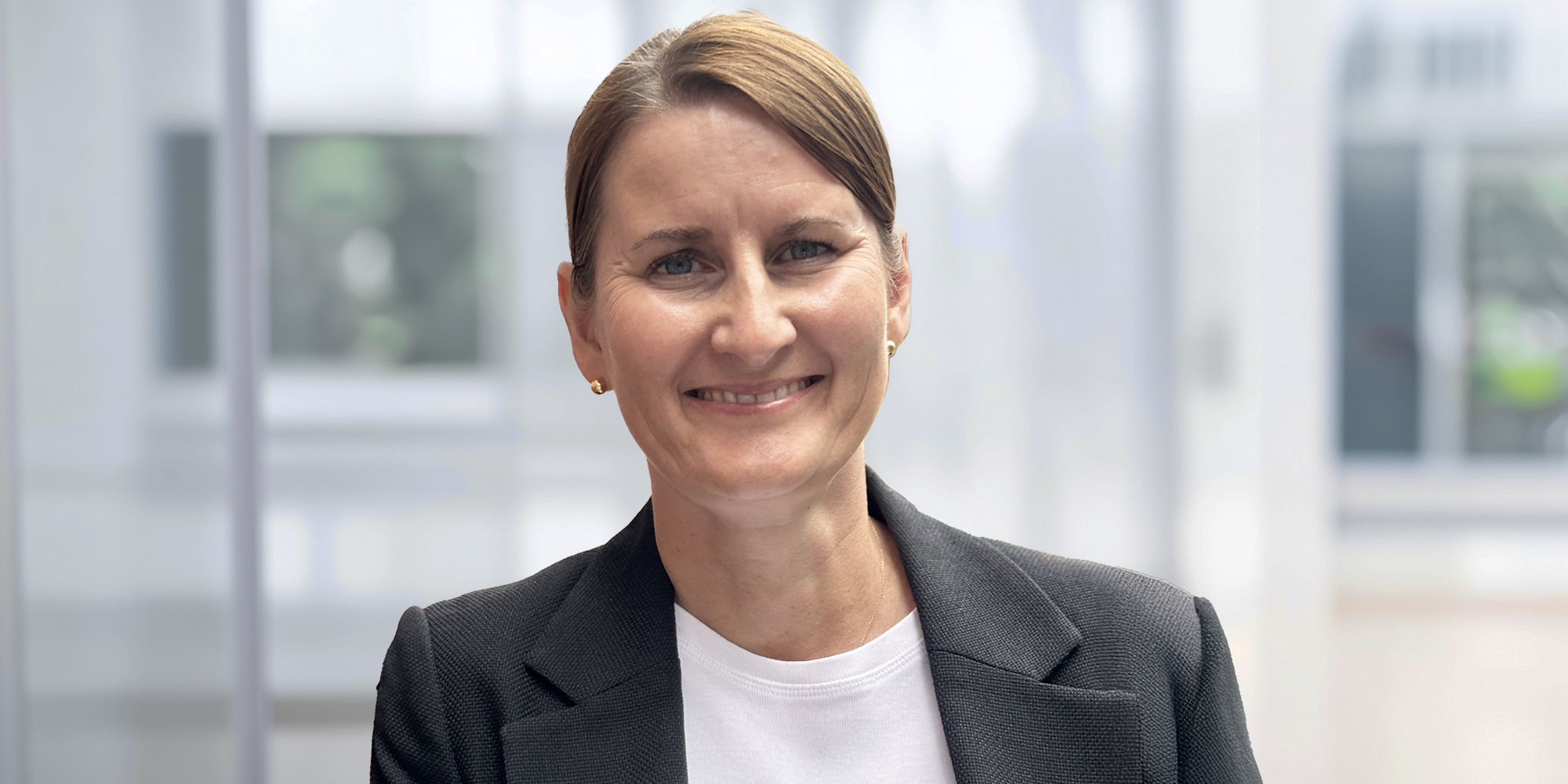 Nancy Schmidt, Mitarbeiterin der Sparkassenversicherung SFG-HT. Porträt einer lächelnden Frau mit zusammengebundenem Haar, in grauem Blazer und weißem Oberteil, vor einem hellen, unscharfen Hintergrund.