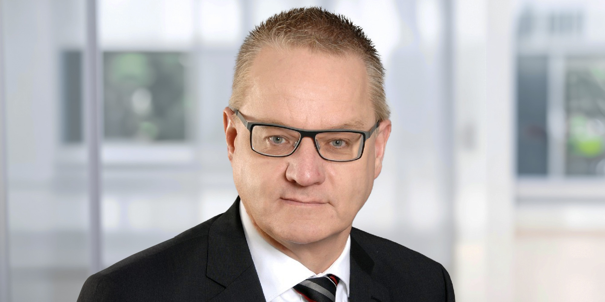 Porträt eines Mannes mit kurzem Haar und Brille, im dunklen Anzug mit Hemd und Krawatte, vor einem hellen, unscharfen Hintergrund.